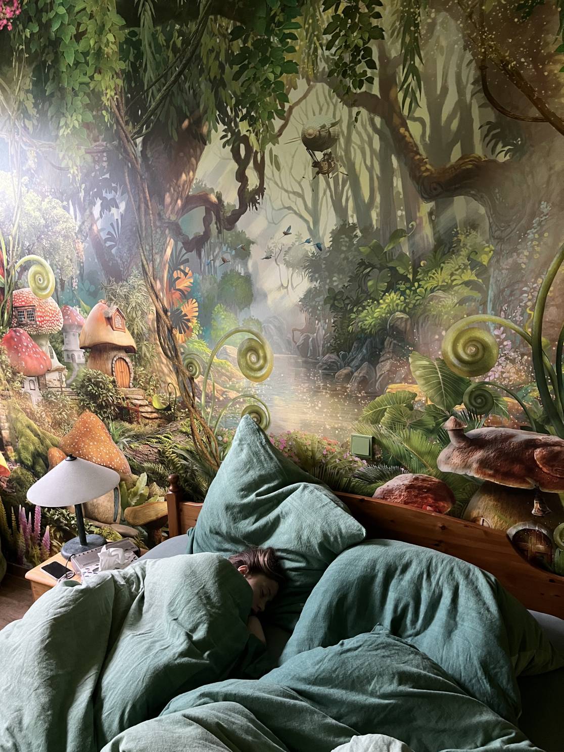 schlafendes Kind unter Bettdecken im Schlafzimmer der Ferienwohnung Alpakahof in Rambow (Plattenburg, Prignitz) mit Blick auf die bemalte Dschungelwand hinter dem Bett mit Wechselschalter und Nachttisch