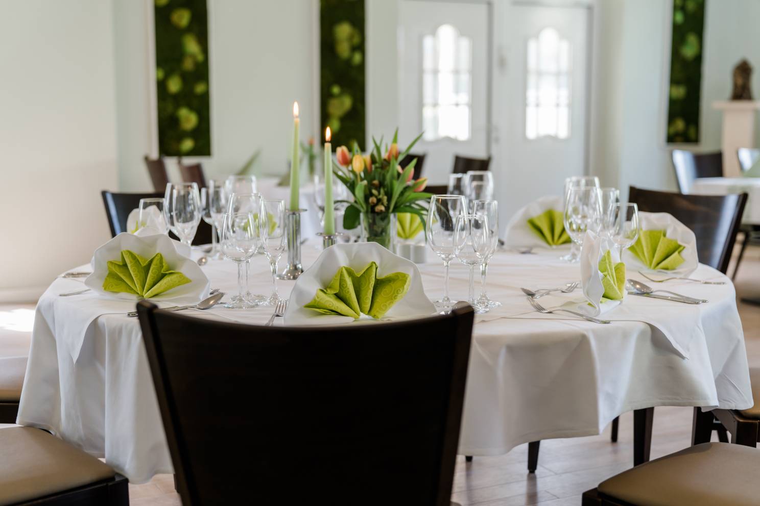 runder Tisch mit dunkeln Stühlen, weiß gedeckt mit grünen Servietten, Gläser und Kerzen, Tulpenstrauß