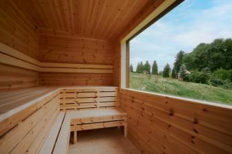 Vorschaubild Innenraum einer Sauna mit Holzbänken mit einem grpßen Fenster und Blick auf Wiese und Bäume sowie hellblauen Himmel