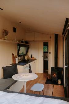 Vorschaubild Innenraum mit hözernem Boden einer Sitzecke mit Bank und Hocker, ein Kamin und eine Küchenzeile mit Blick auf die Badkabine