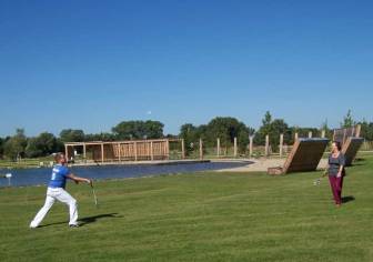 Vorschaubild Paar spielt am Teich im neuen Kurpark von Bad Wilsnack auf einer gemähten sattgrünen Wiese Federball, im Hintergrund Holzliegen und ein Steg zum Pausieren und Flanieren
