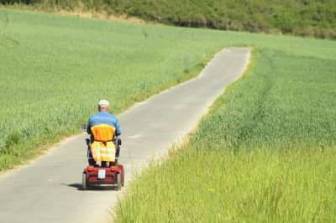Vorschaubild Mann mit blauem Pullover und beigefarbener Mütze fährt mit Elektro Rollstuhl mit Warnweste auf einen aspahltierten Weg entlang eines im Sommer gut bewachsenen Feldes