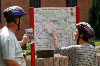 Vorschaubild zwei Radfahrer stehen mit einer Karte vor der Leistungsträgertafel sowie der Karte zum Knotenpunktradeln in der Prignitz im Europäischen Storchendorf Rühstädt im Biosphärenreservat Flusslandschaft Elbe