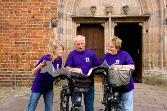 Vorschaubild drei Radfahrer studieren mit ihren Rädern vor der roten Ziegelwand und den Türen der Wunderblutkirche Bad Wilsnack eine Radfahrkarte und unterhalten sich lächelnd
