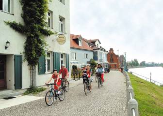 Vorschaubild Familie radelt auf dem Elbdeich entlang des Elberadweges an der Elbe vorbei an Zimmervermietungen und Restaurants in Wittenberge im Sommer