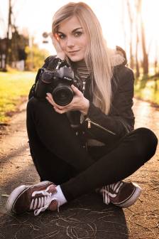 Vorschaubild lächelnde Mitarbeiterin Monique Bessert mit blonden langen Haaren, schwarzer Jacke, schwarzer Jeans und Turnschuhen sitzt im Schneidersitz auf dem Asphalt im Sonnenuntergang und hält eine Nikon-Kamera in der Hand