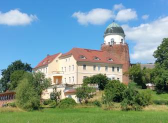 Vorschaubild historische Burg Lenzen, in der sich heute ein Restaurant und ein Hotel befinden, sowie der Burgturm, in dem sich ein Museum und eine Ausstellung vom BUND befinden, vom Burgpark aus im Sommer unter strahlend blauem Himmel