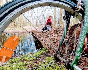 Vorschaubild durch Speichen des Fahrradreifens fotografierte Radfahrerin, die auf einem Baumstamm, der quer über einen kleinen Wasserlauf liegt, im Schneidersitz eine Pause macht 