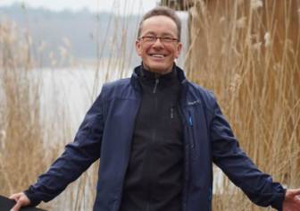 Vorschaubild Natur- und Landschaftsfüher Jürgen Paul steht vor einem See mit Schilf, lachender Mann mit ausgebreiteten Armen in Funktionskleider mit braunem lockigen Haar und Brille