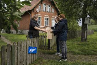 Vorschaubild Frîa Hagen, Inhaberin des Café Elbeglück in Mödlich überreicht lachend am Zaun ihres Gartenlokals einer Mutter mit ihrem jugendlichen Sohn zwei Lunchtüten "Radlerparadies Prignitz"