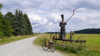 Vorschaubild Elberadweg, Elbdeich im Sommer mit Fahrrad neben blühender Wieser, auf der ein mystischer Fährmann, der sogenannte "Eiserne Charon", eine Figur vom in dem Ort Mödlich ansässigen Künstler Bernd Streiet, steht