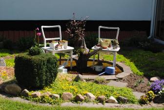 Vorschaubild Zum Tag der Offenen Gärten öffnet der Garten der Familie Schneider mit dekorativen Stühlen auf denen kleine Pflanztöpfe stehen sowie einem von einer Steinpackung umfassten Blumenbeet