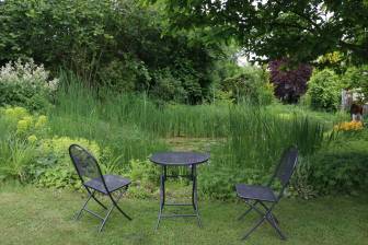 Vorschaubild Zum Tag der Offenen Gärten öffnet der Garten der Familie Tolksdorf mit zwei Stühlen und einem Tisch an einem Teich mit üppig grüner Bepflanzung, an dem Besucher entlang spazieren