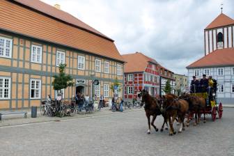 Vorschaubild vierspännige Postkutsche mit uniformierten Kutschern auf dem Marktplatz von Wusterhausen Dosse inmitten von Fachwerkhäusern, u. a. dem Herbst'schen Haus, in dem sich das Wegemuseum befindet und vor dem Menschen die Kutsche bewundern