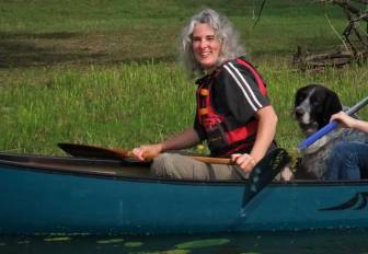 Vorschaubild die zertifizierte Natur- und Landschaftsführerin Susanne Figueiredo sitzt mit ihrem Hund in einem grünen Kanu mit einem Paddel, lächelnde Frau mit grauem Haar und einer roten Schwimmweste