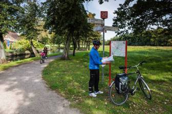 Vorschaubild Radfahrer studiert eine Radkarte neben seinem Fahrrad vor einem Knotenpunkt mit Wegweisern und Leistungsträgertafel im Park Perleberger Hagen an der Stepenitz