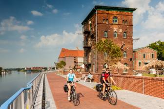 Vorschaubild Paar Radfahrer auf der Elbepromenade und dem Elberadweg in Wittenberge in der Kulisse der historischen Speicher des Elbe Resort Alte Ölmühle, neben dem sich heute eine Strandbar mit Blick auf die Elbe befindet