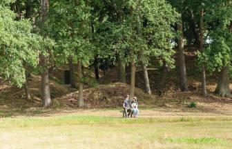 Vorschaubild Pärchen mit Hüten und Sonnenbrillen sitzen auf einer steinernen Bank vor dem Wald und Hügel, hinter dem sich das Königsgrab Seddin versteckt
